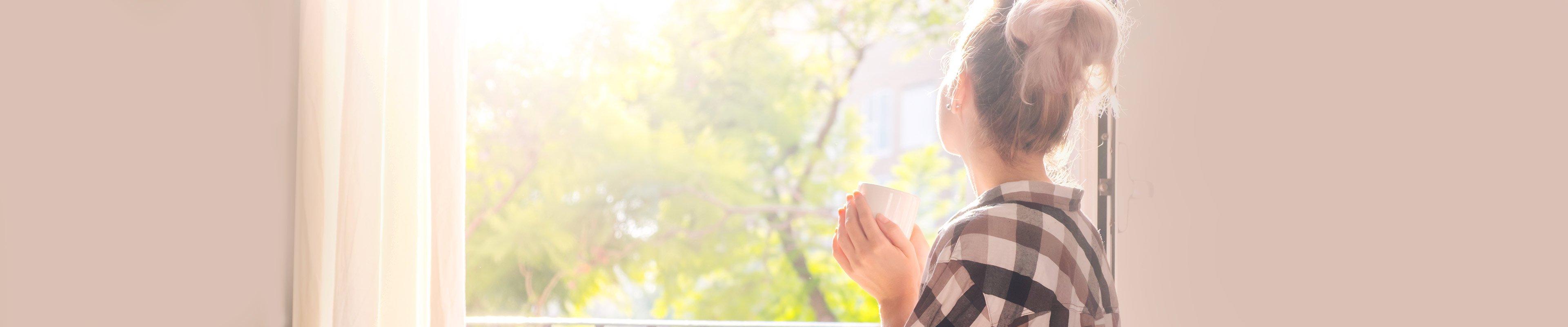 Woman standing at window drinking coffee. 